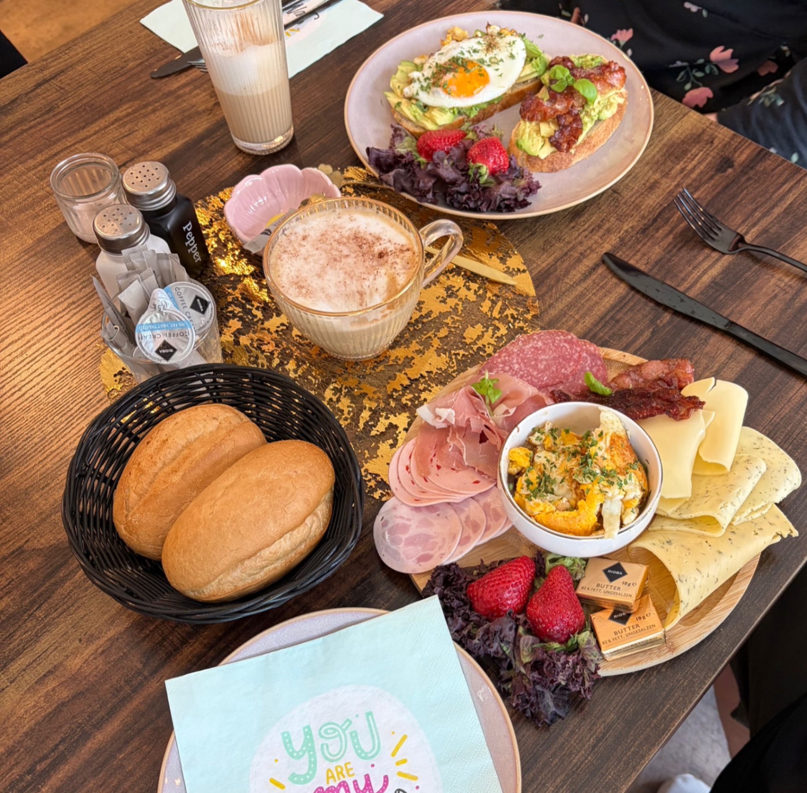 Frühstücksspread mit Brötchen, Wurst, Käse, Rührei, Avocado-Toast und Cappuccino